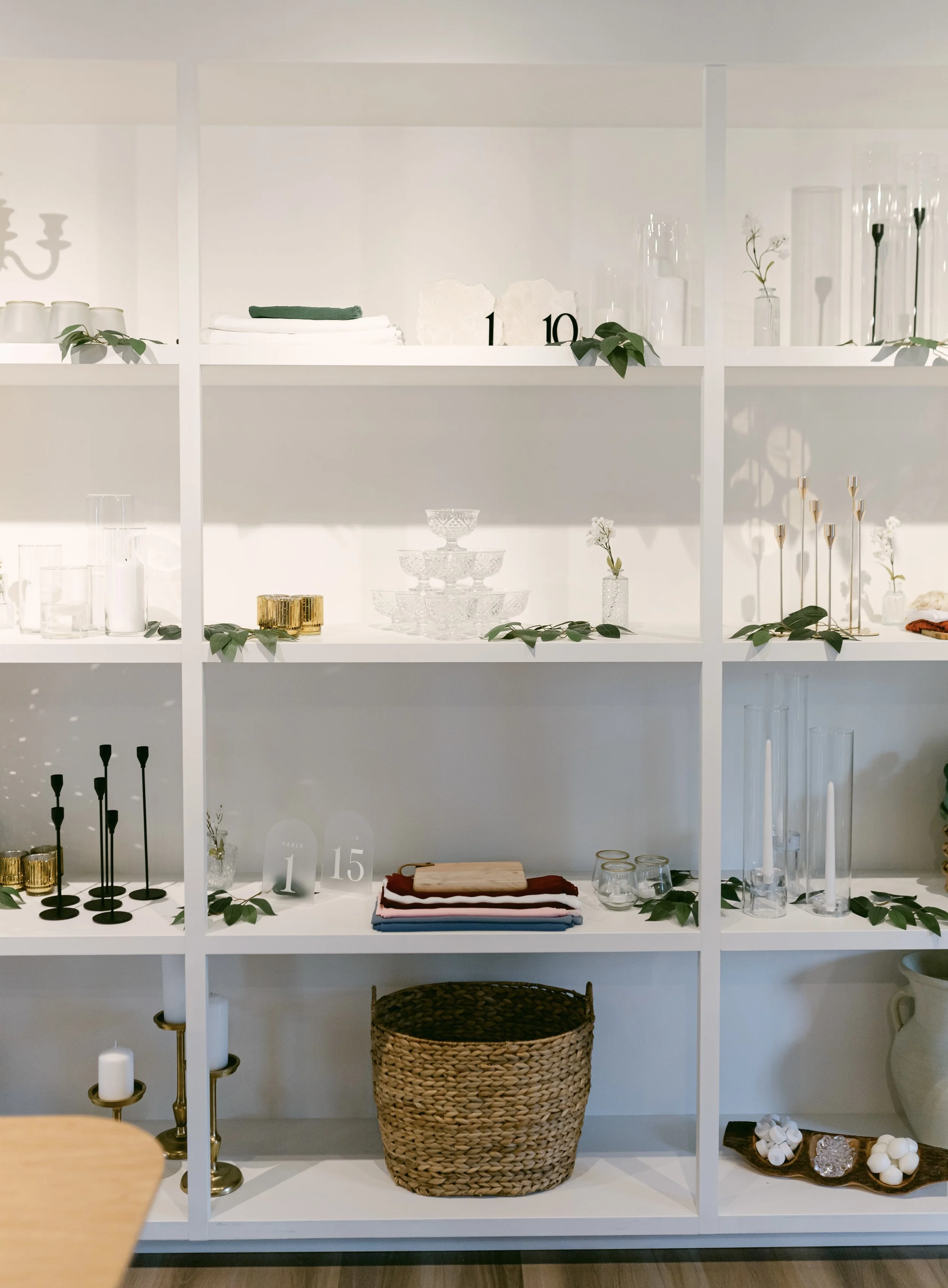 A white shelving unit with decorative items including candles, vases with flowers, glassware, a wicker basket, folded textiles, candle holders, and table number signs.