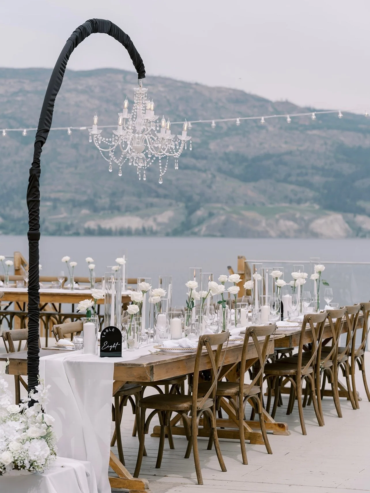Outdoor wedding or event setup with long wooden tables decorated with white flowers, candles, and glassware. A black chandelier hangs above, and a scenic water and mountain view in the background.