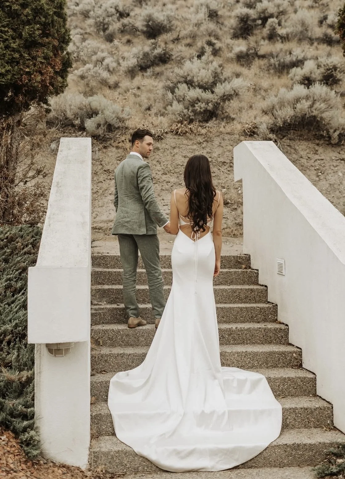 A couple in wedding attire holding hands on outdoor stairs with a desert landscape background.