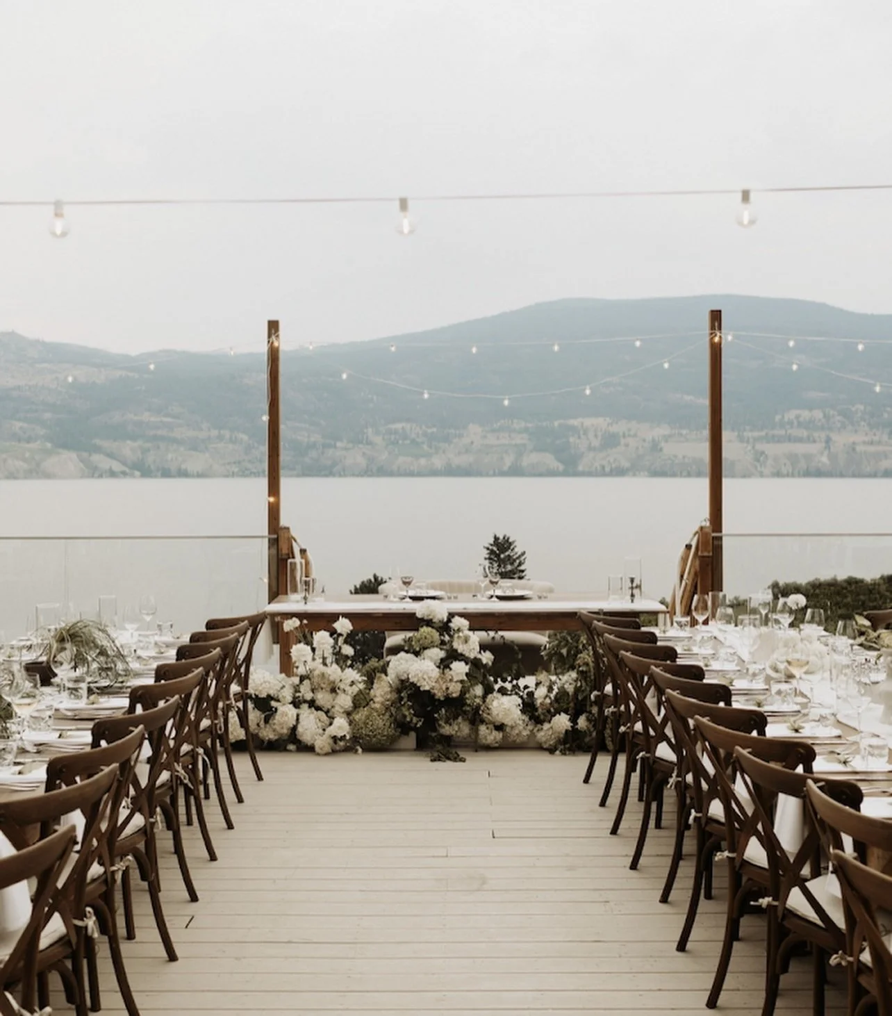 Outdoor wedding reception setup with a long table decorated with white flowers and place settings, overlooking a lake with mountains in the background, and string lights overhead.