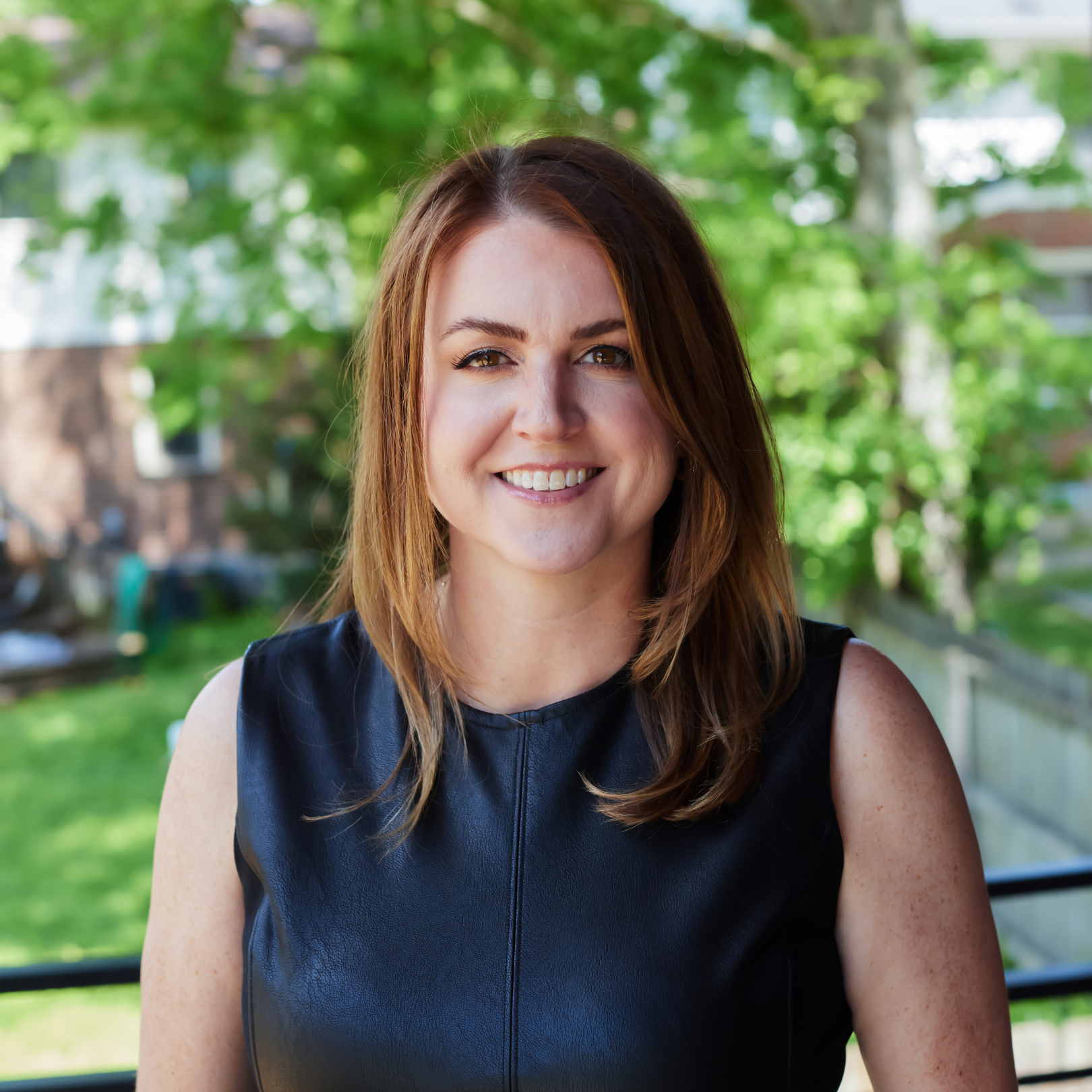Real estate broker and developer Erin Tunney of The Tunney Group, with long auburn hair and light skin, smiling outdoors against a green, leafy background.