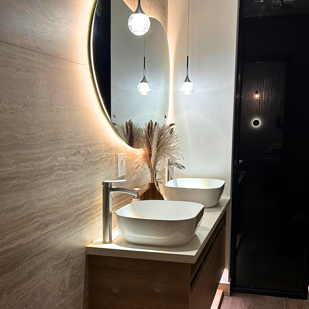 Modern bathroom vanity with two white vessel sinks, a round backlit mirror, a vase with pampas grass, and hanging pendant lights reflected in the mirror.