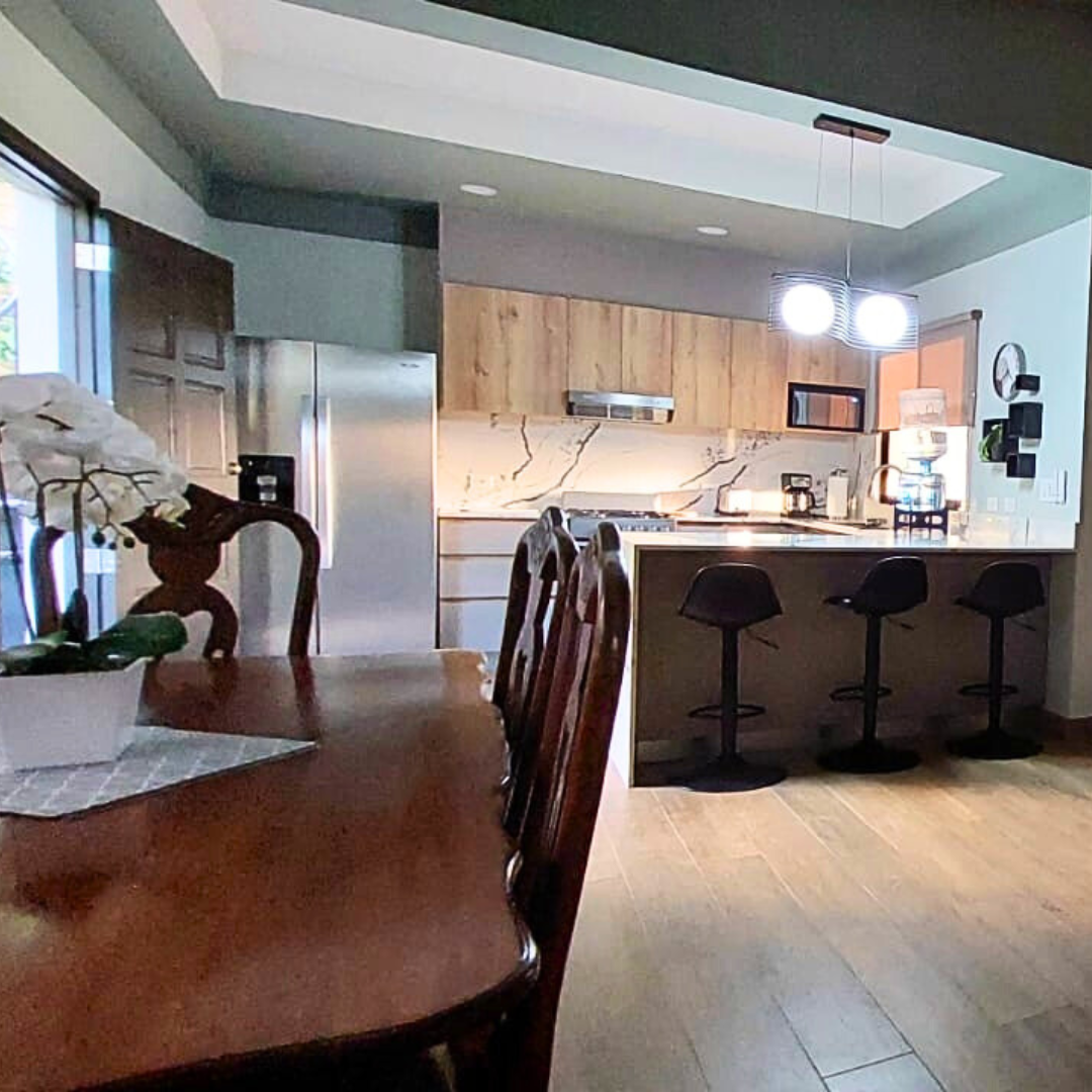 A modern kitchen with wooden cabinets, a marble backsplash, a stainless steel refrigerator, and a breakfast bar with three black stools. A dining table with chairs is in the foreground, and there are decorative items and appliances on the countertops