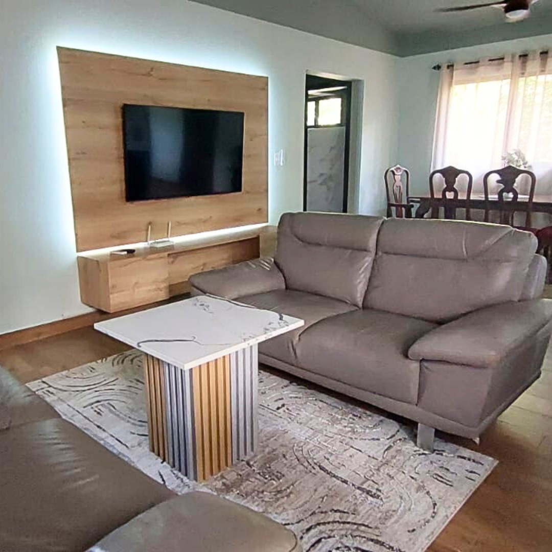 Living room with a gray sofa, a wooden TV wall with a mounted flat-screen TV, a white coffee table with wood trim, a patterned rug, and a dining area with a dark wooden table and chairs near a window with curtains.