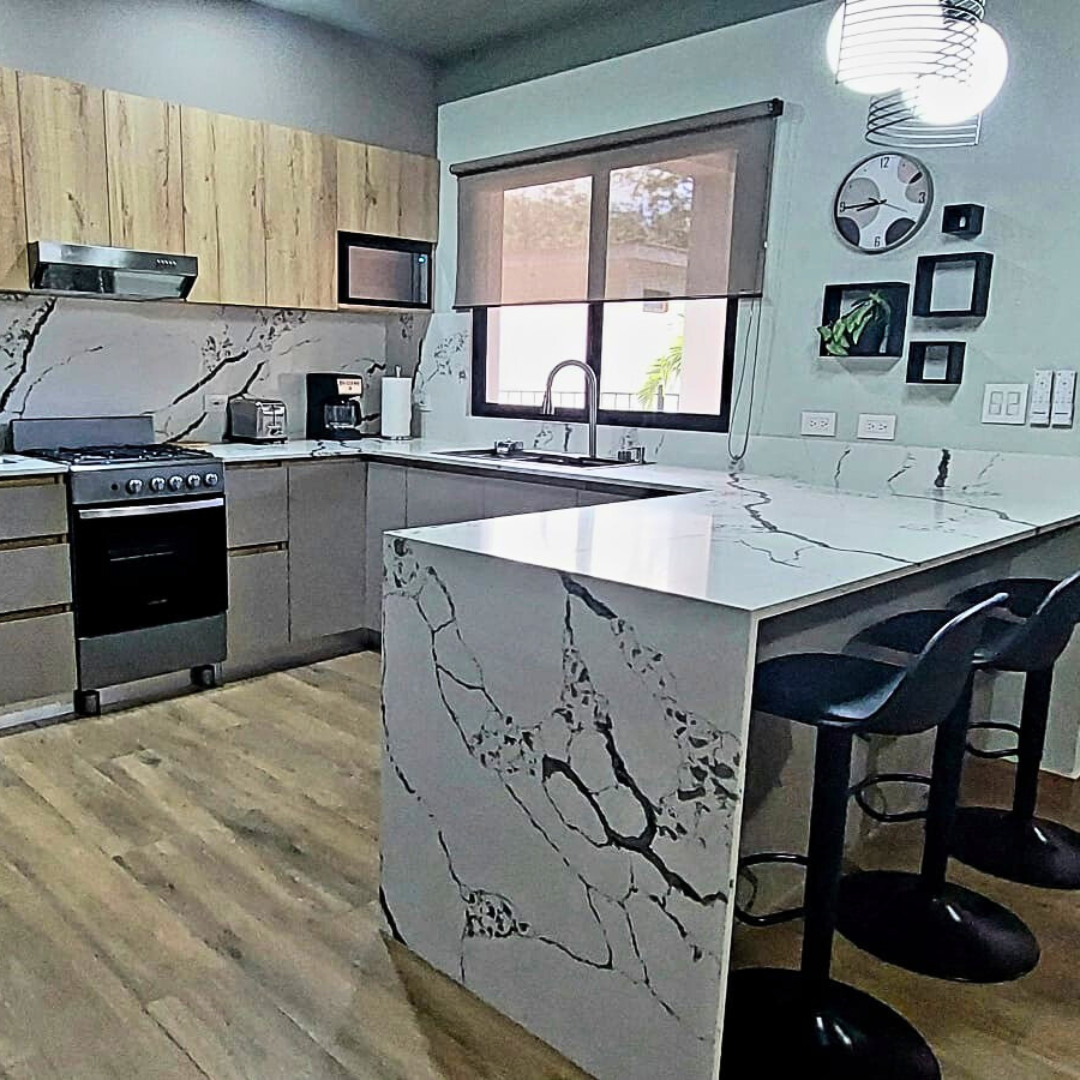Modern kitchen with marble countertops and backsplash, wooden upper cabinets, stainless steel appliances, a window with roller shade, black bar stools at a marble island, and decorative wall art.