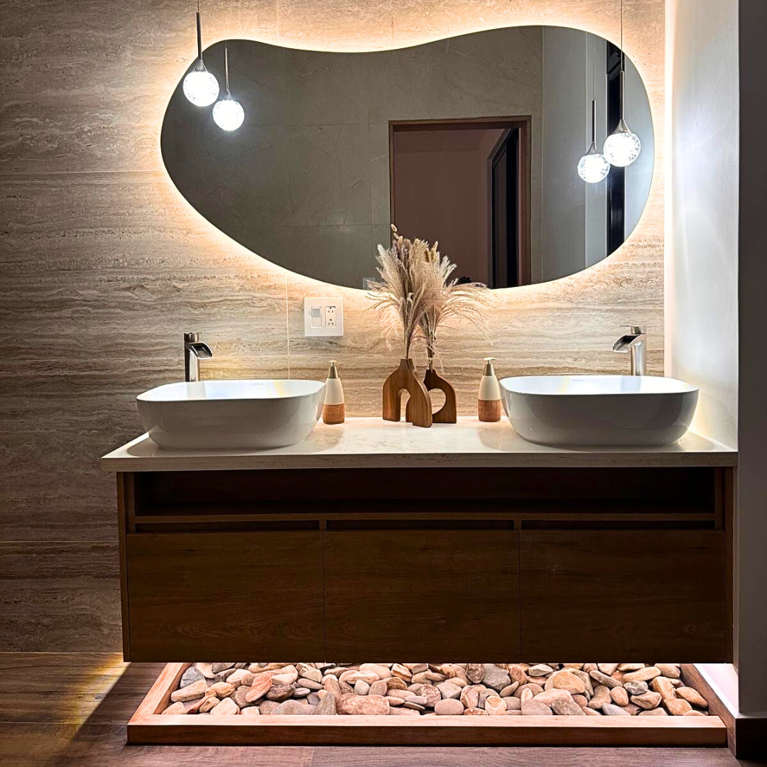 Bathroom vanity with two vessel sinks, large oval backlit mirror, decorative vases with dried plants, and pebble flooring under the vanity.