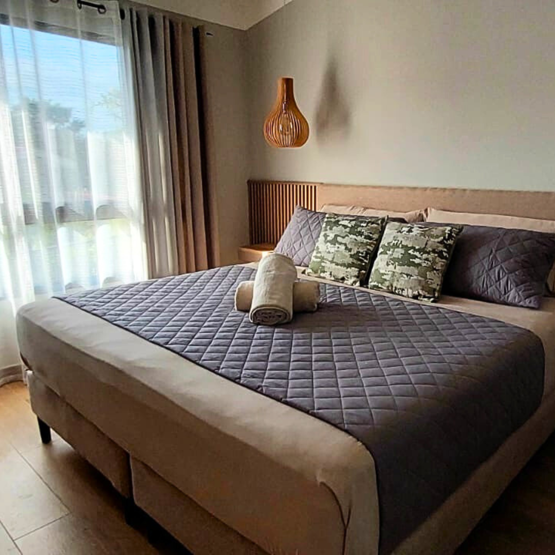 A neatly made bed with gray quilted bedding, decorative pillows, and two rolled towels on top, situated in a bedroom with large window and beige curtains.