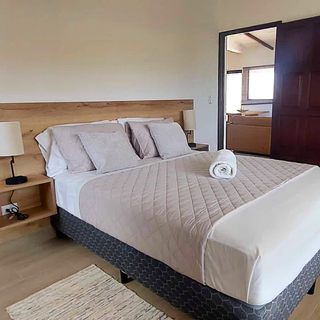 A neatly made bed with a light beige quilt and four pillows, a rolled white towel on top, two nightstands with lamps, and a window in the background showing a bathroom with a bathtub.