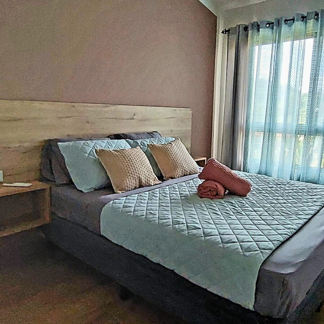 A bedroom with a bed featuring beige pillows, blue pillows, and a quilted white blanket. There are rolled orange towels on the bed. The room has a pink accent wall, a window with sheer and dark curtains, and a wooden nightstand.