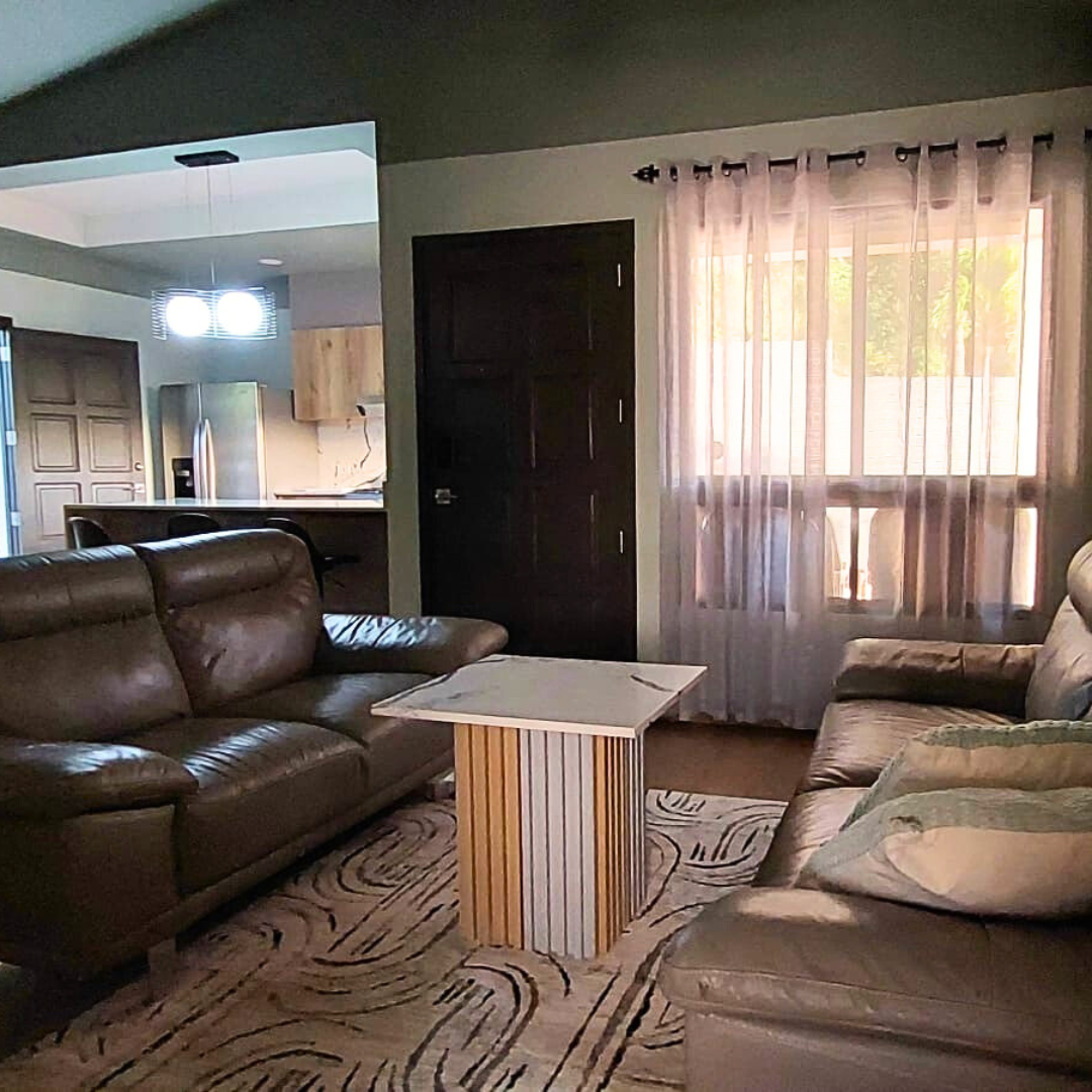 Living room with two brown leather sofas, a white and wood coffee table, a window with sheer curtains, and a view into the kitchen area with a refrigerator and overhead lighting.