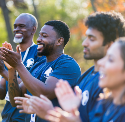 Group of diverse people wearing blue shirts clapping and smiling outdoors in a park with autumn foliage.
