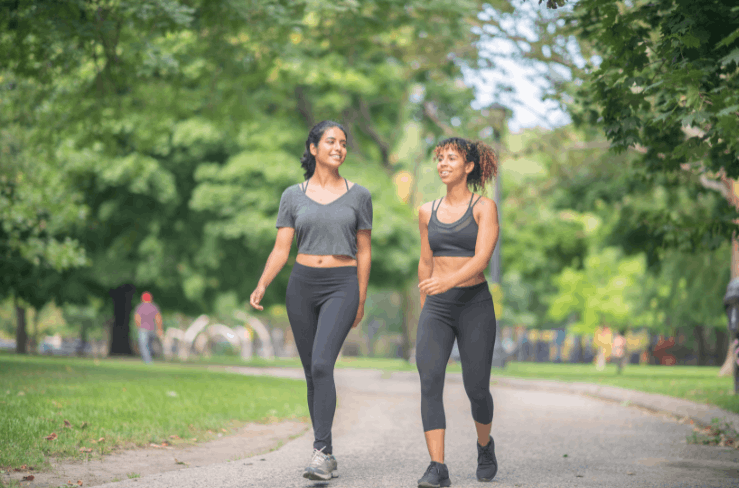 Two women walking and talking outdoors on a grassy park trail, surrounded by green trees and other park visitors.