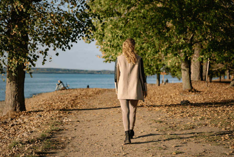 Woman walking along a leaf-covered path by a lake, surrounded by trees with green leaves during fall.