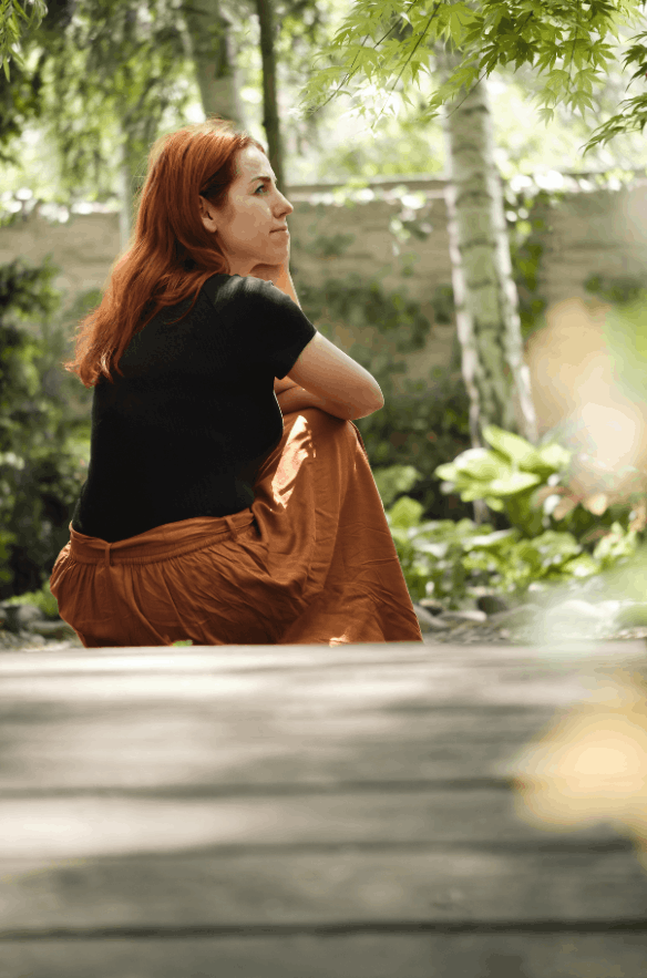 A woman with red hair sitting outdoors in a garden with trees and plants, resting her chin on her hand and gazing to the side.