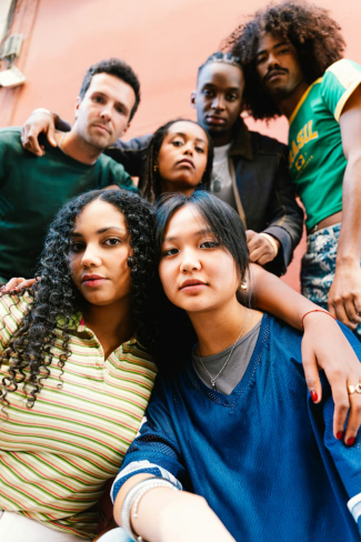 A diverse group of six young adults posing together outdoors during daytime, with a pink building in the background.