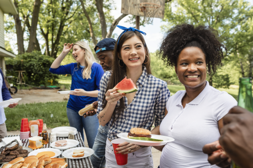 Group of diverse friends enjoying a backyard picnic with food and drinks, smiling and chatting outdoors.