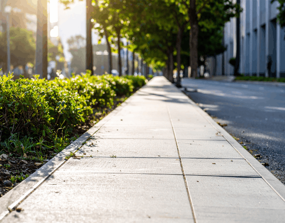 Sidewalk with green bushes and trees lining the sidewalk on one side and a street on the other, with modern buildings in the background