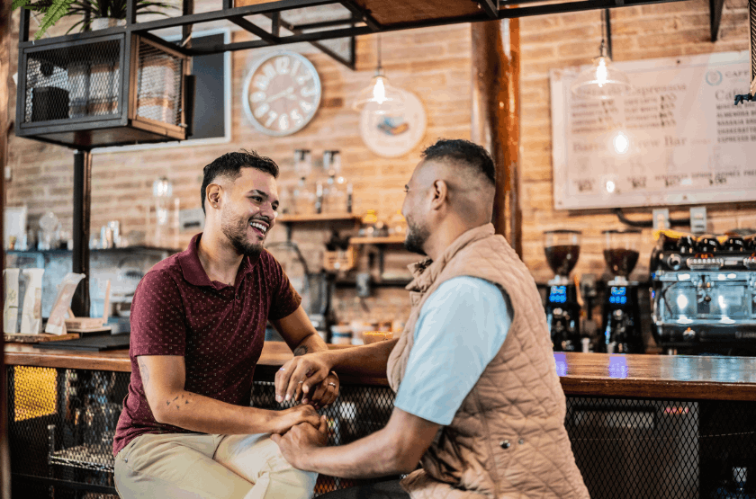 Two men are smiling and talking at a bar counter in a cafe, holding hands.