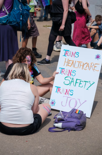 A person sitting on the ground with a transgender pride flag, writing or drawing next to a sign that promotes trans awareness and rights, with other people walking in the background.