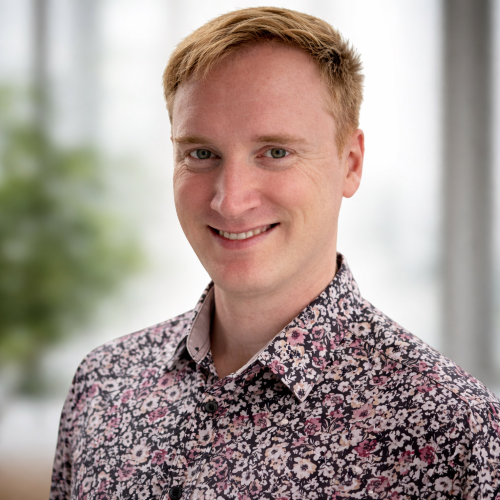 A smiling man with short, light brown hair wearing a patterned shirt in front of a blurred background with greenery.