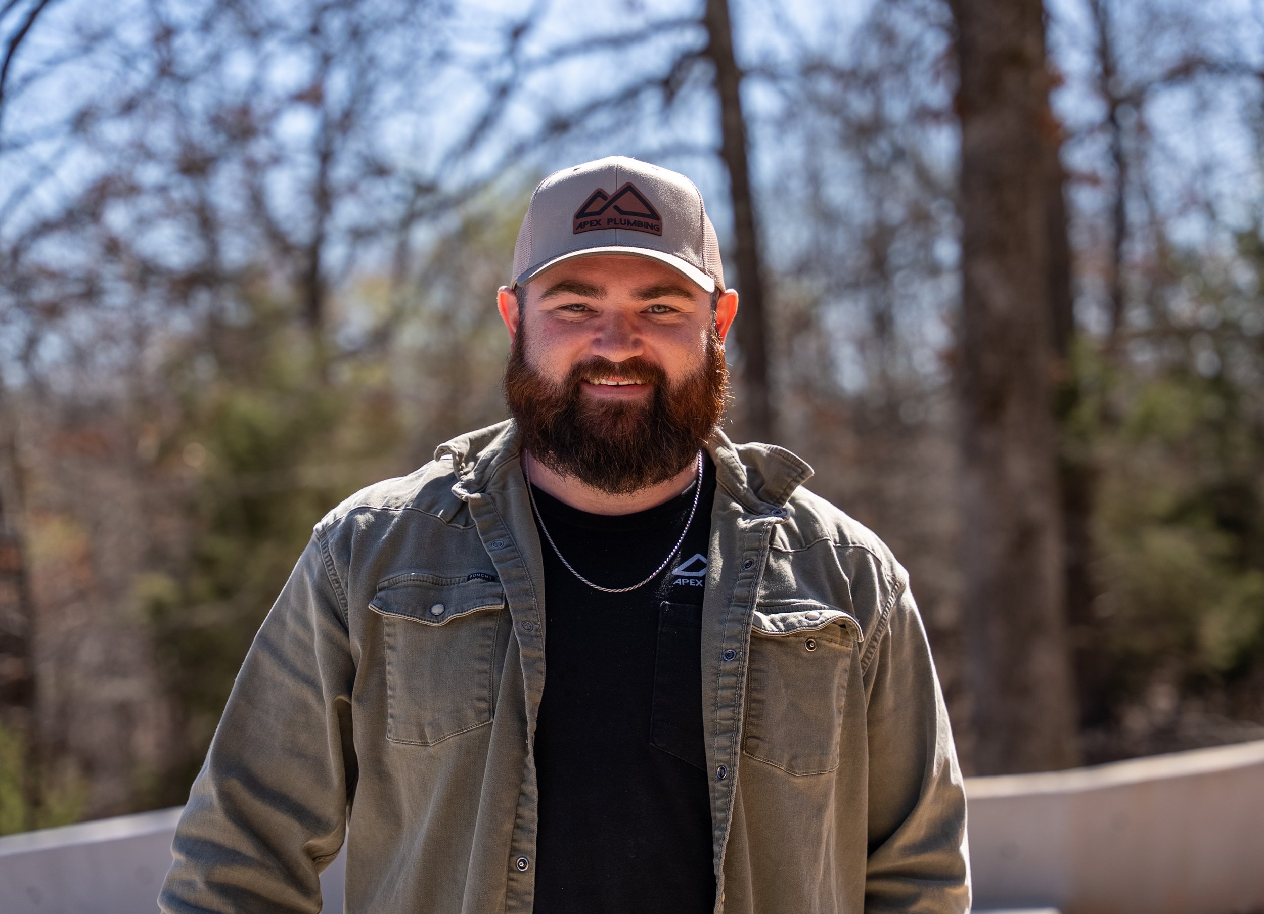 A bearded man wearing a gray Cap with a logo that reads 'APEX PLUMBING' and a gray jacket, smiling outdoors with a background of leafless trees and a blue sky.