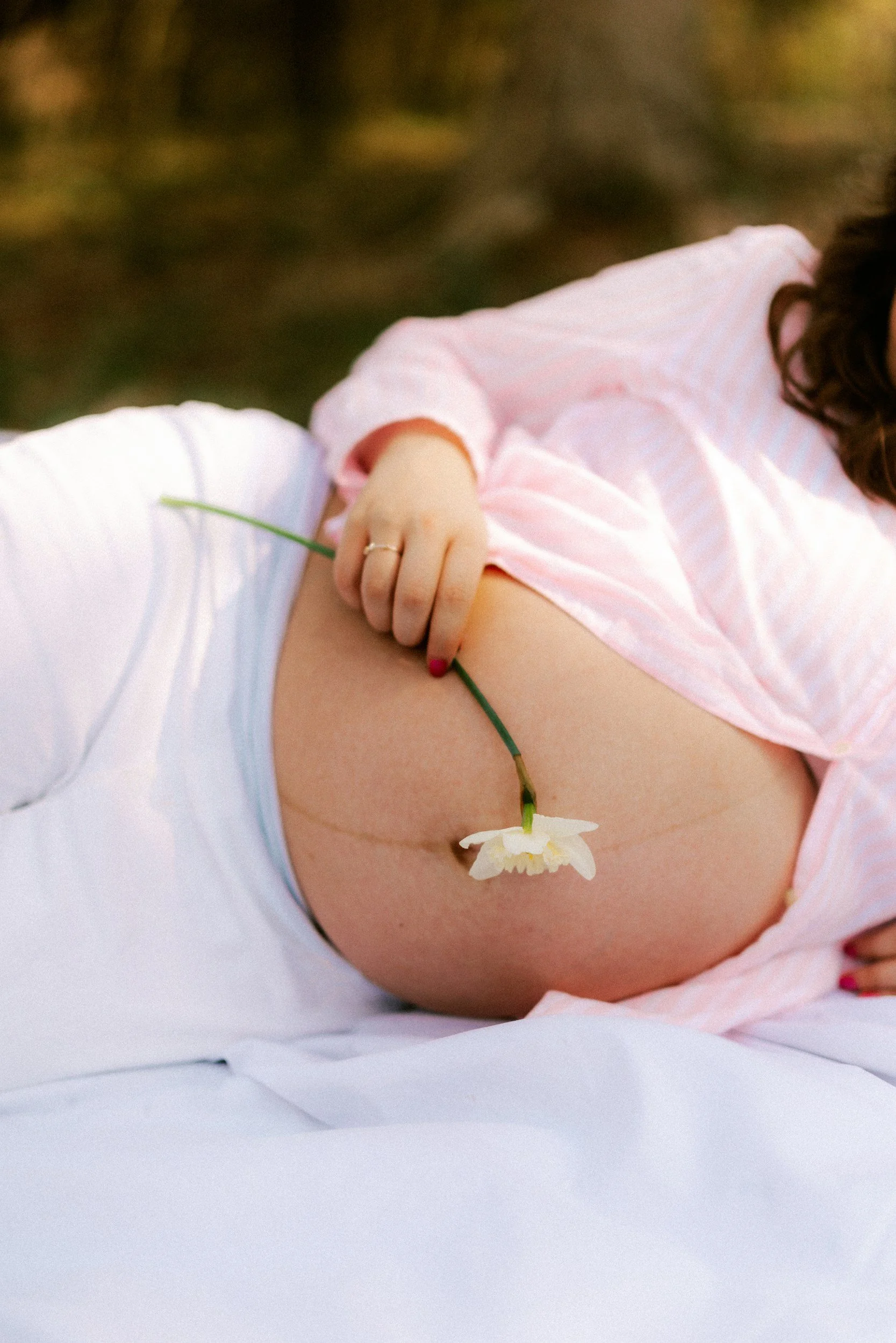 A pregnant person in pink pajamas lying on a bed holding a white flower, with part of their arm and hand visible. Pregnancy health and support.