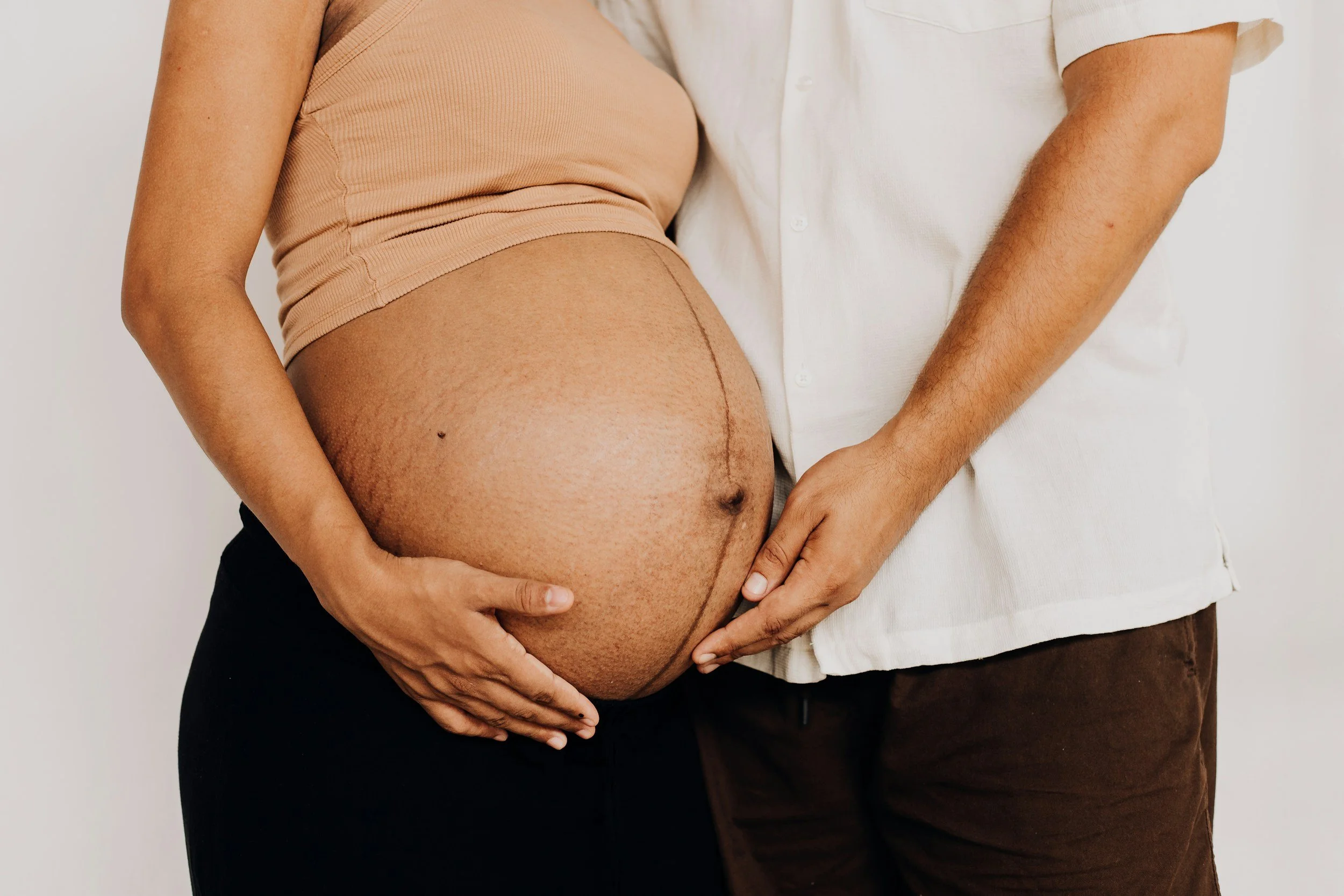 A pregnant woman and a man holding her belly and waist against a plain background.