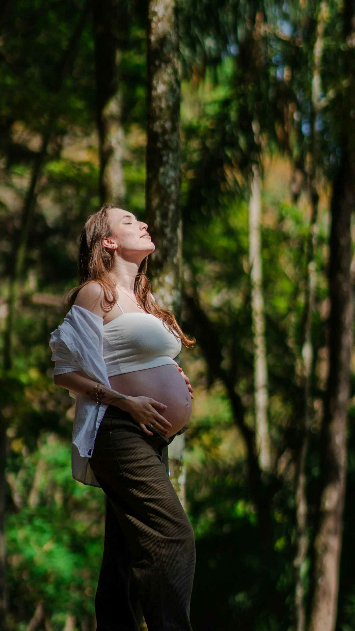 A pregnant woman stands outdoors in a wooded area, with her hands on her belly, enjoying the sunlight with her eyes closed.