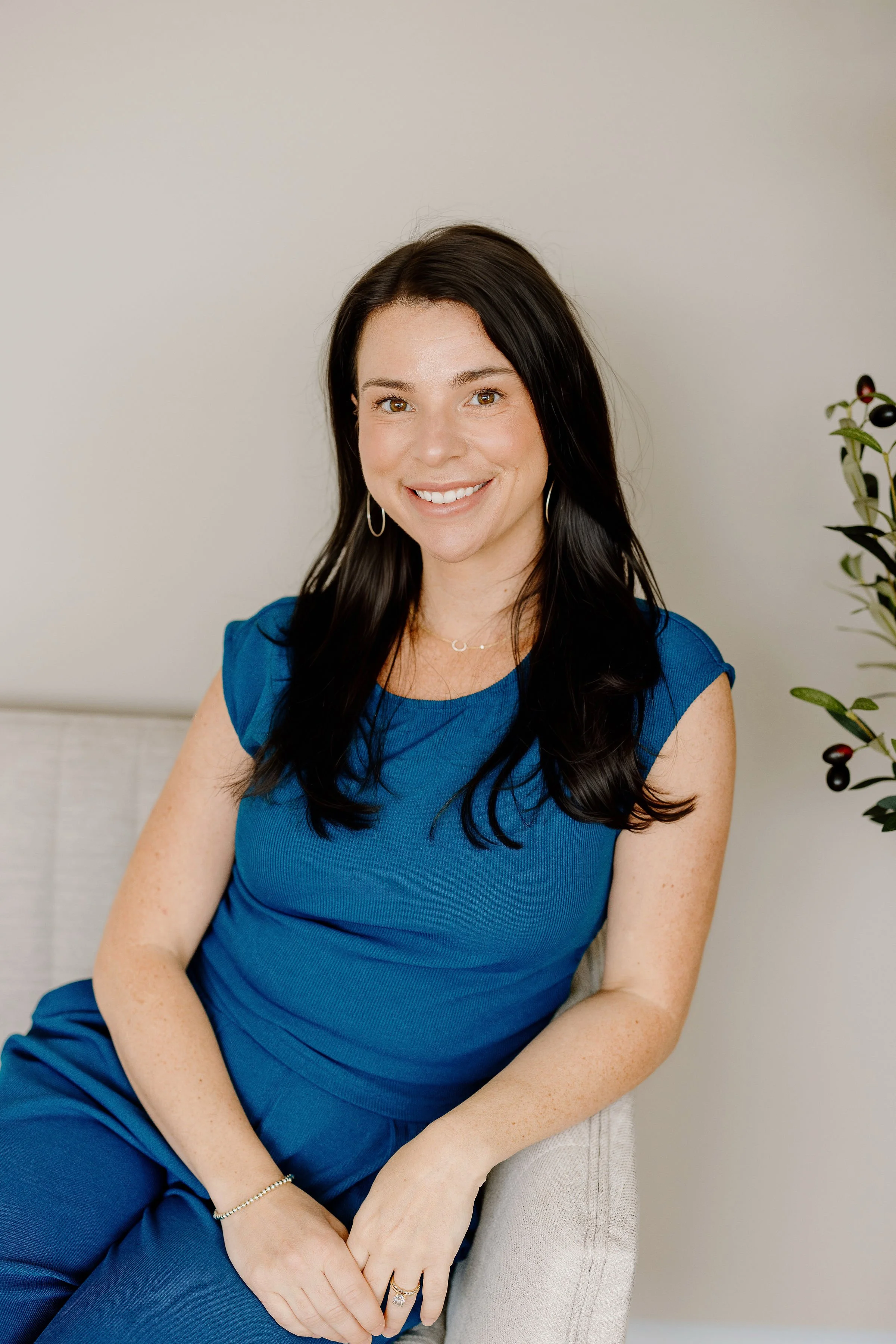 A woman, Maryland Acupuncturist and Birth Doula, with long dark hair, brown eyes, and a bright smile, sitting on a light-colored chair against a plain light background, wearing a blue sleeveless dress and gold jewelry.
