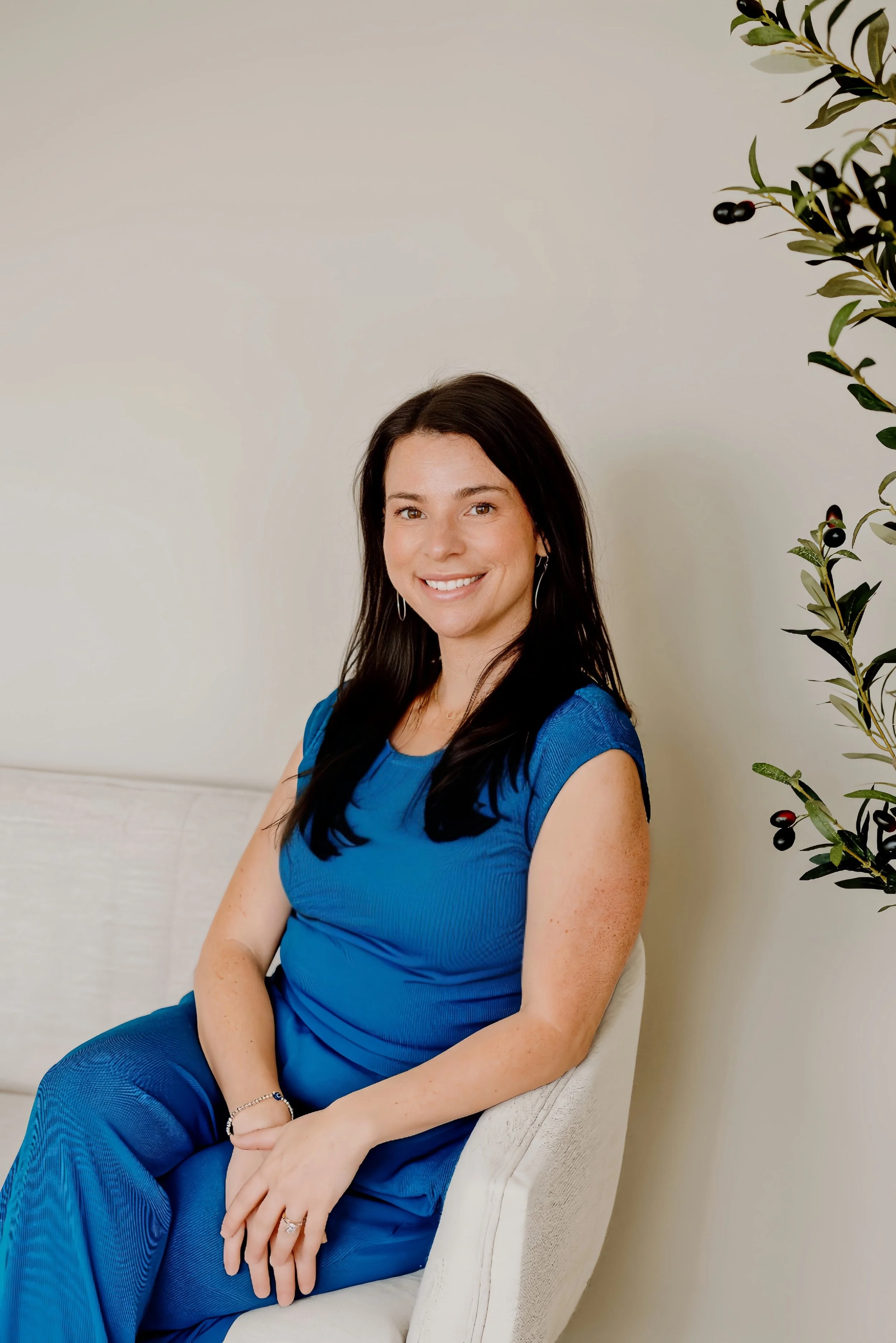A woman, Maryland Acupuncturist and Birth Doula, with long dark hair, wearing a blue dress, sitting on a white chair, smiling, with a beige wall and a potted plant with black berries to her right.