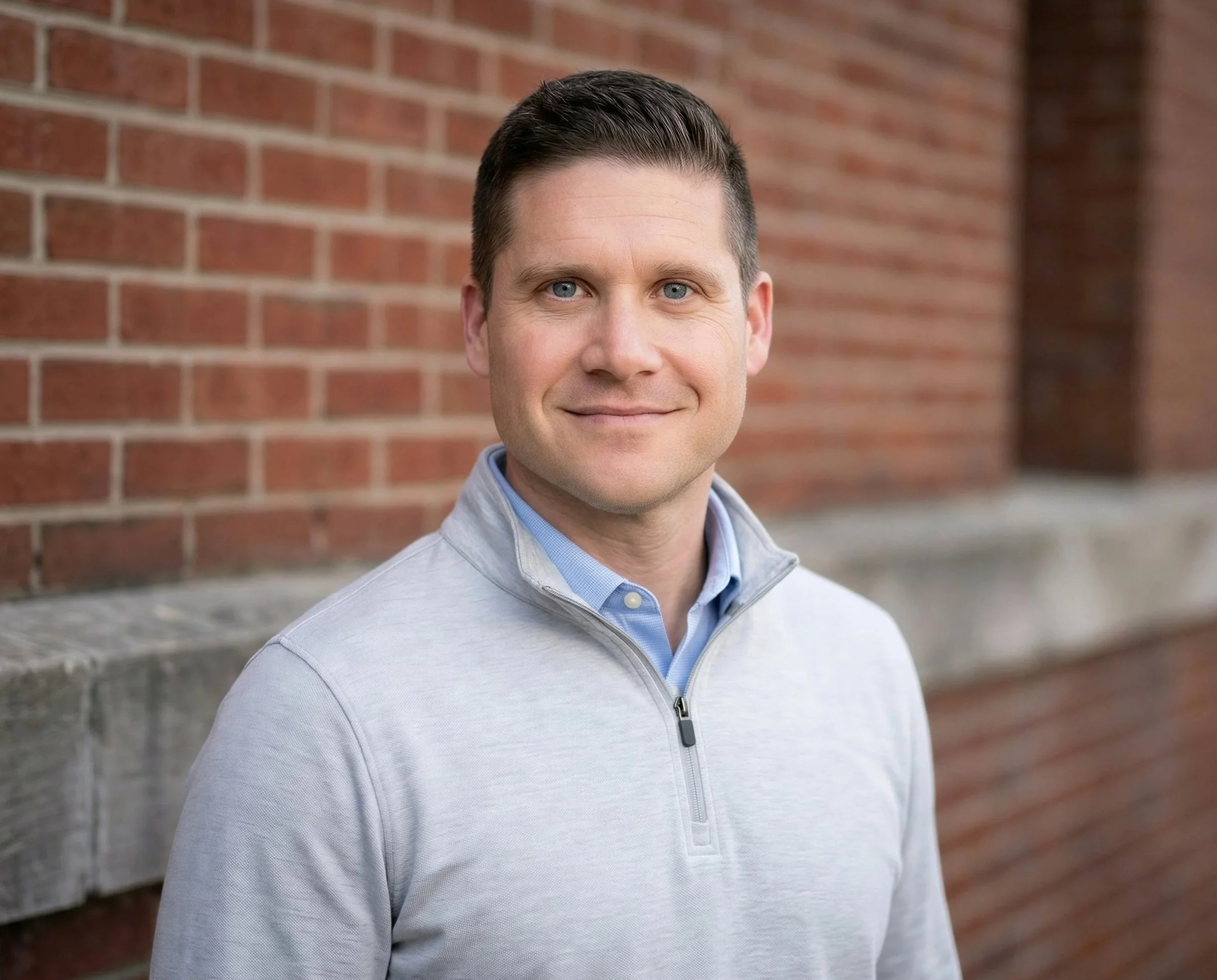 A man with short dark hair, blue eyes, and wearing a light gray quarter-zip sweater with a blue collared shirt underneath, standing outdoors in front of a brick wall, smiling at the camera.