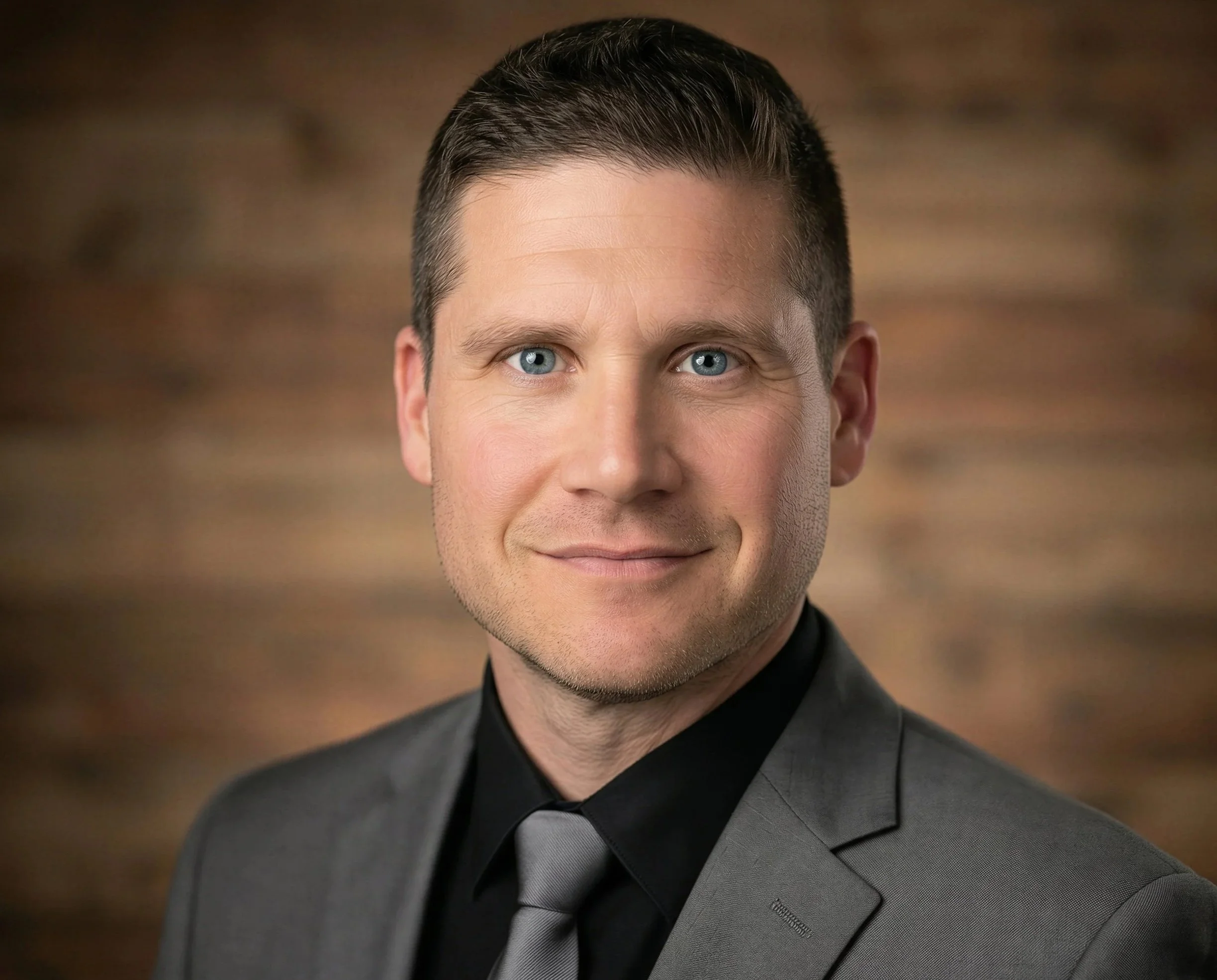 A man in a gray suit, black shirt, and gray tie smiling with a wooden background.