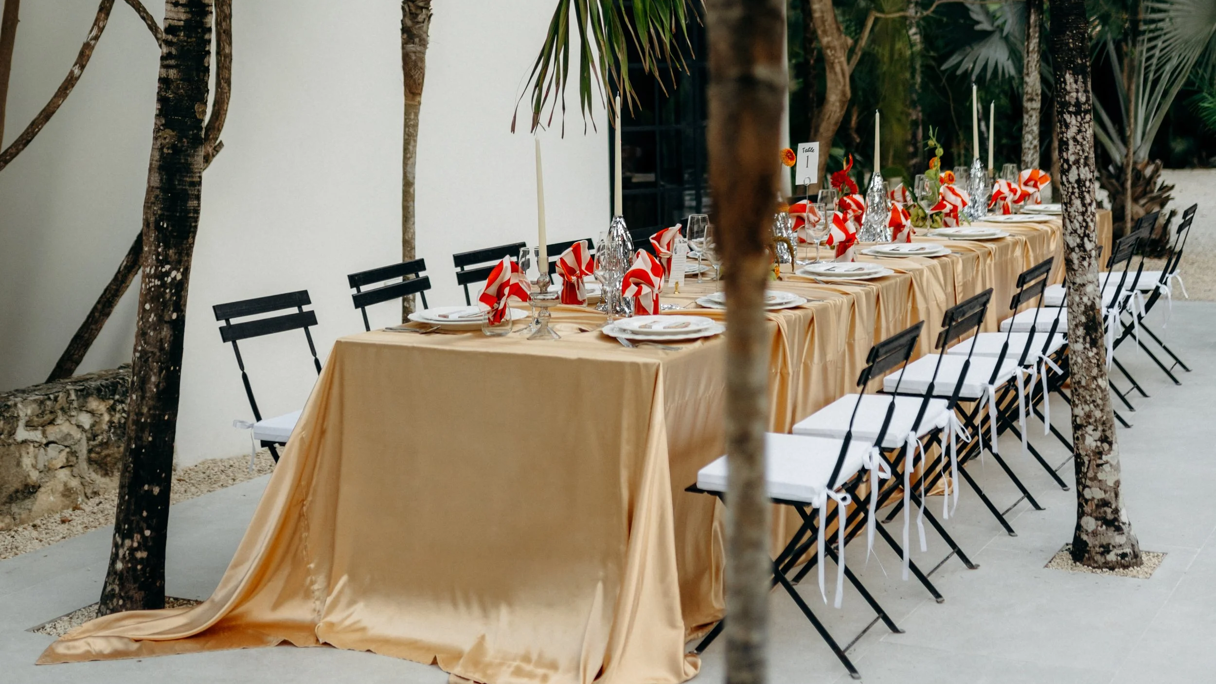 Long rectangular outdoor dining table with a gold tablecloth, set for a formal company event with red and white napkins, glasses, and plates, surrounded by black chairs with white cushions, located under trees with a white wall in the background.