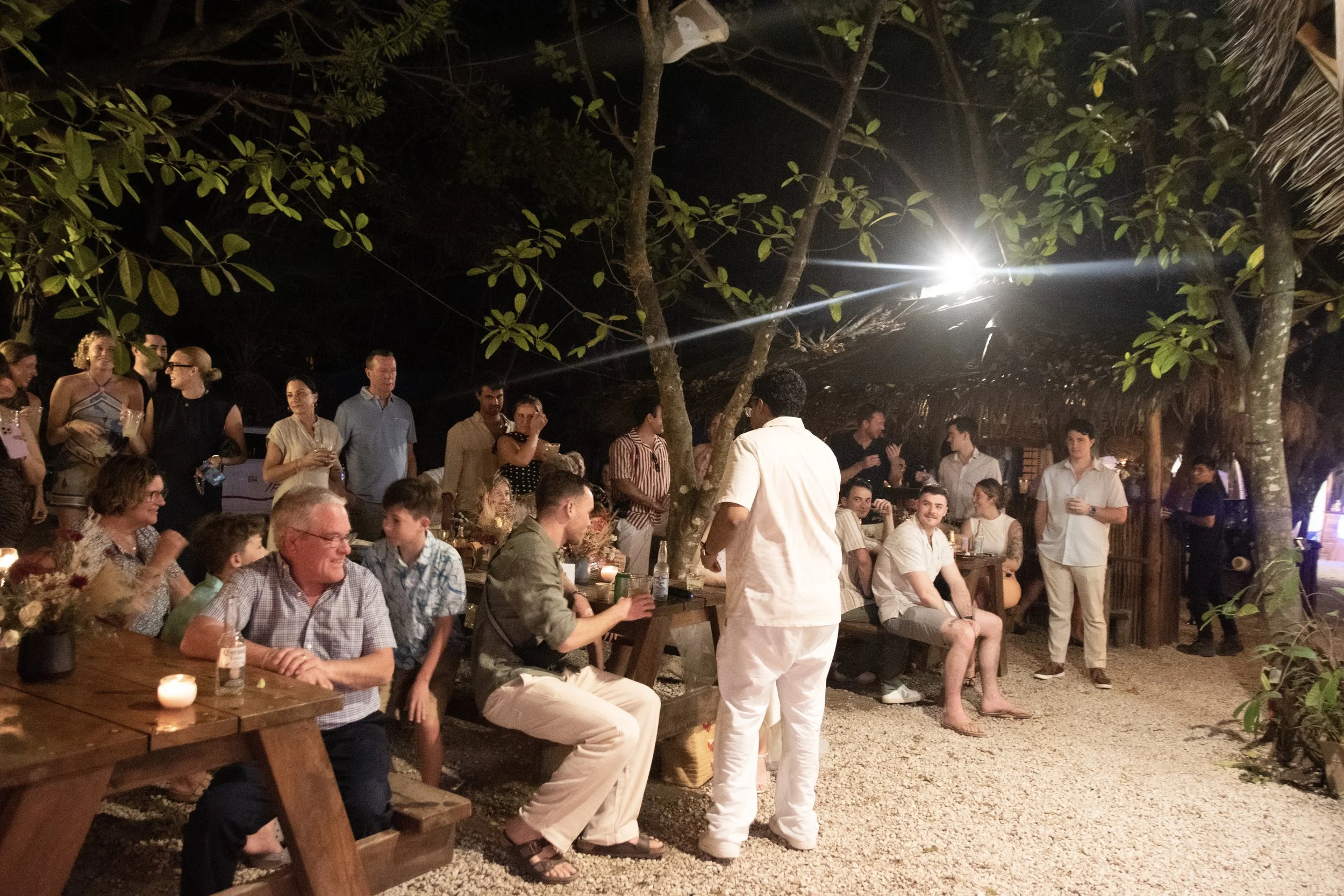 A picture of a launch event party for a startup that was outdoors and shows tables, candles, and string lights, surrounded by trees and tropical foliage, with some individuals standing and others sitting.