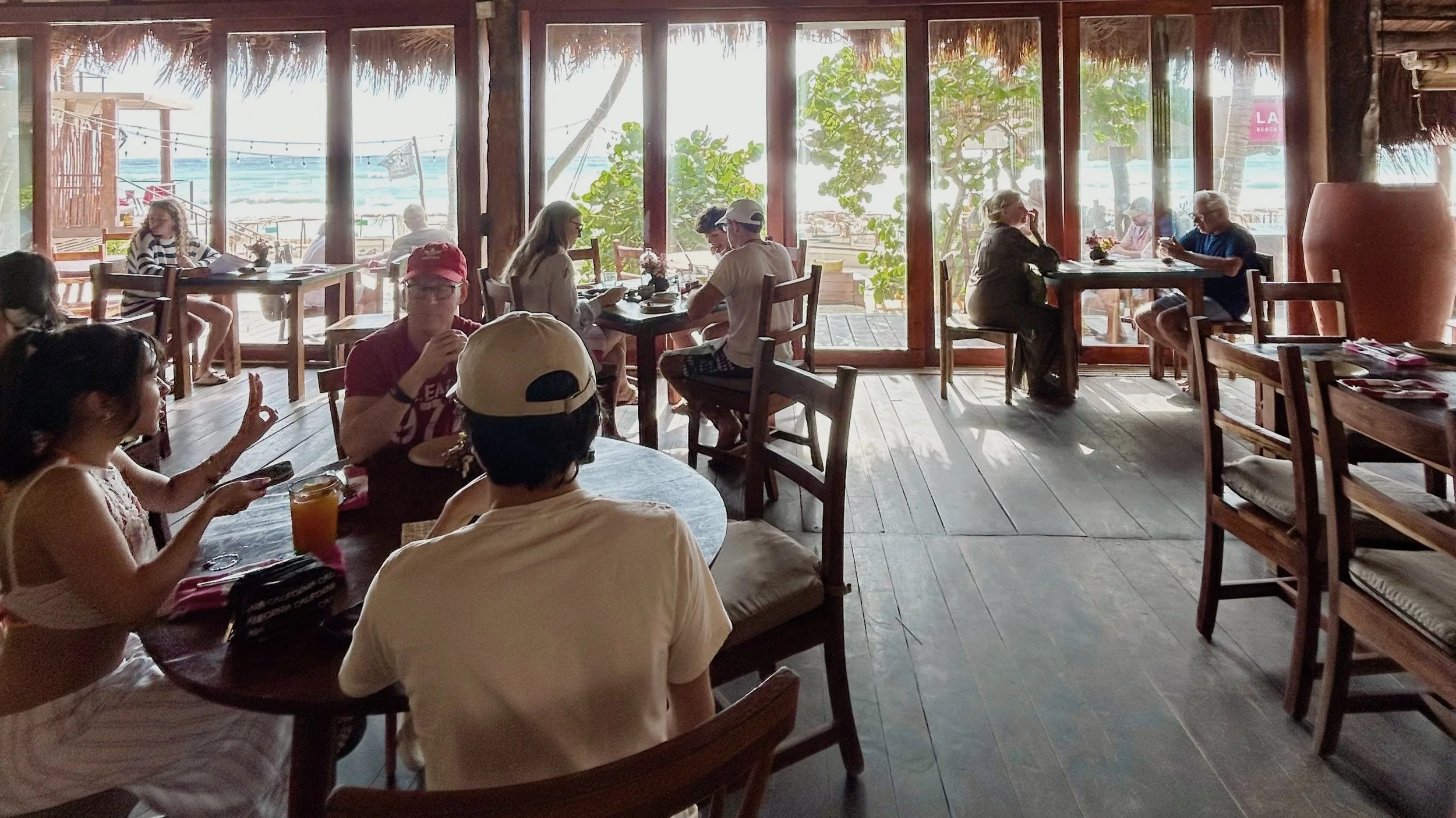 A seaside restaurant with large windows overlooking the beach and ocean, filled with people dining and socializing during a company trip in Tulum, Mexico.