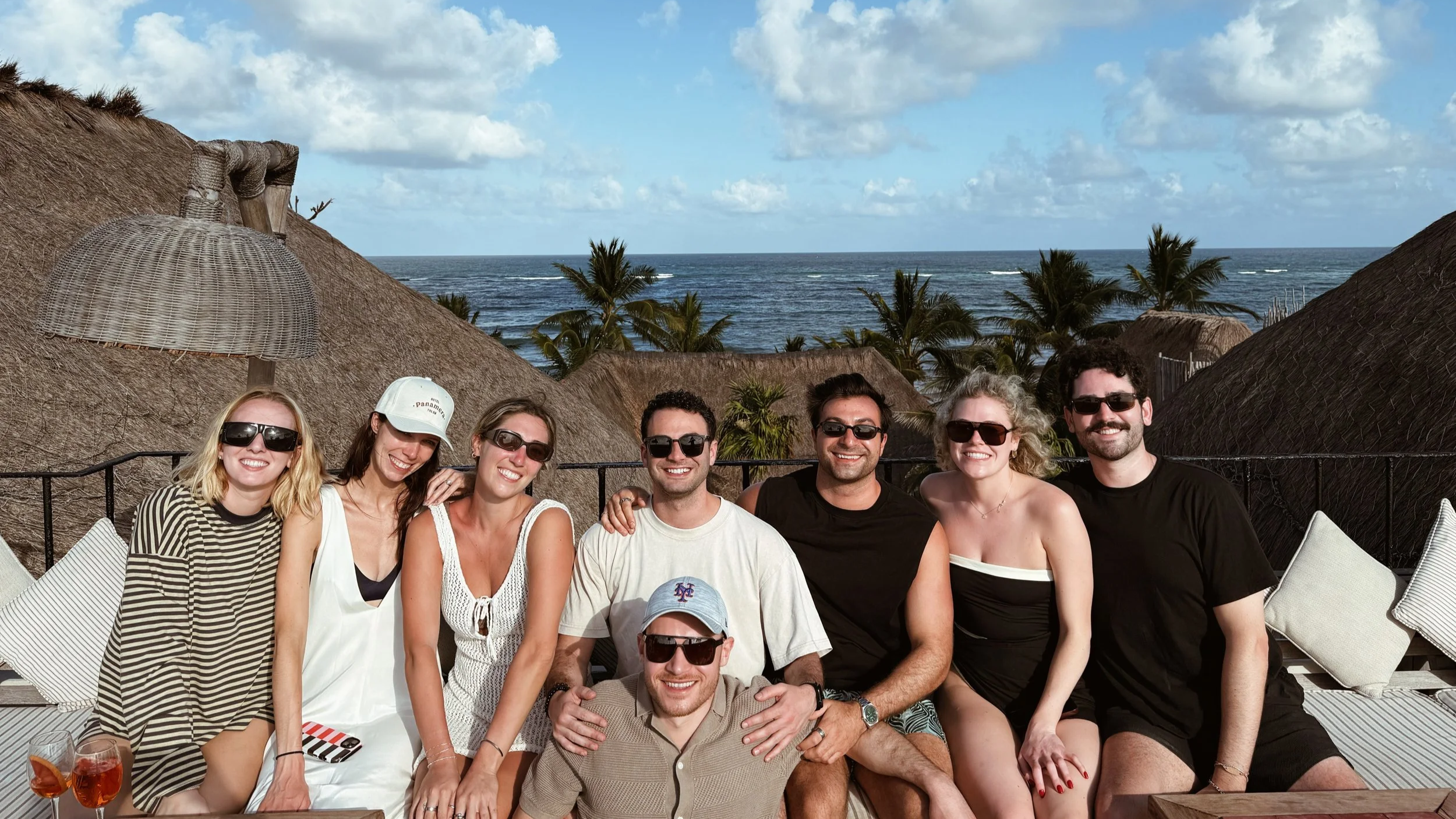 Group of eight friends and colleagues sitting together, smiling, with the ocean, palm trees, and thatched roofs in the background, enjoying sunny weather at their team bonding retreat in Tulum, Mexico.