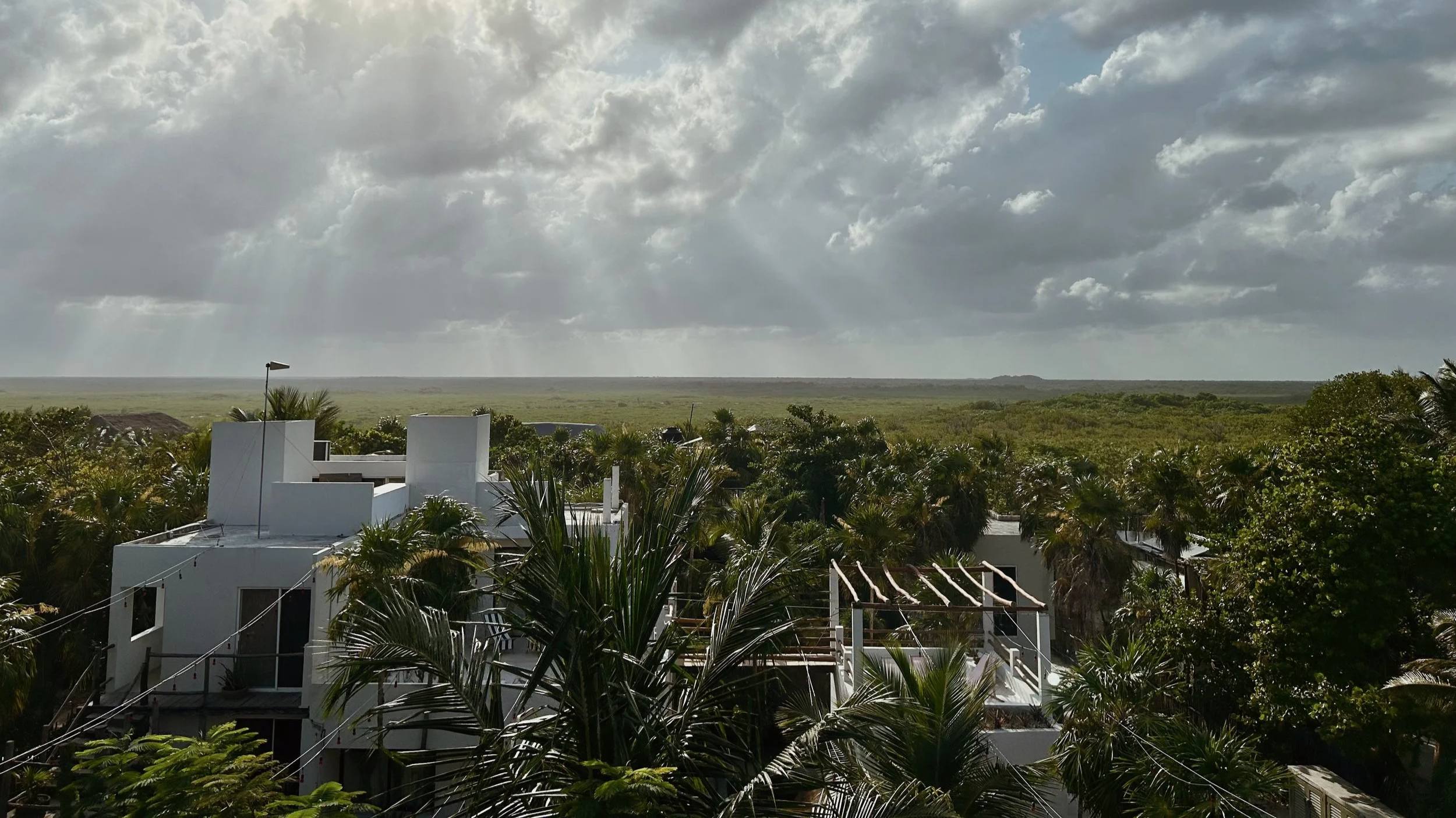 Luxury hotels in Tulum, Mexico surrounded by lush green palm trees and vegetation under a cloudy sky with sunlight rays breaking through