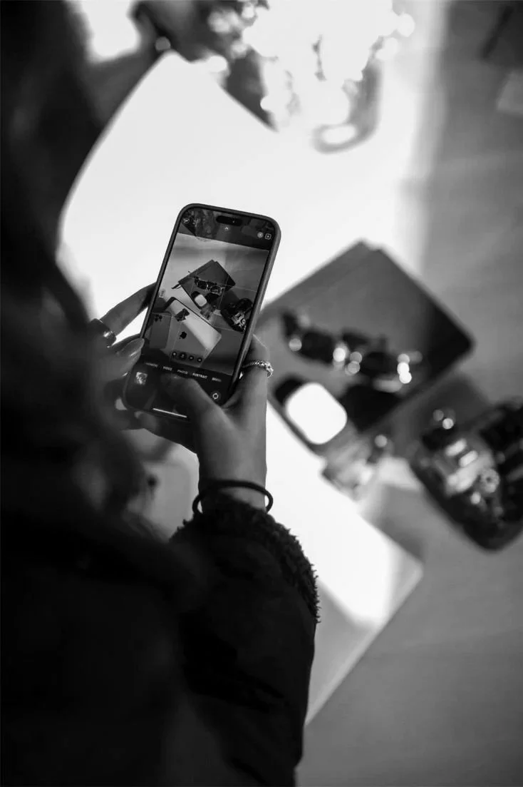 A person taking a photo of a display with a smartphone, showing various objects including sunglasses and a small box, on a table.