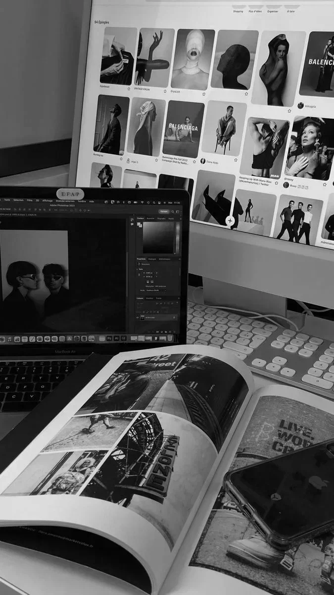 A workspace with two computer screens displaying graphic and fashion images, an open magazine showing photographs and text, a smartphone, and a white keyboard.
