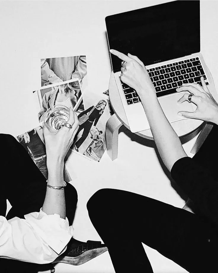 Two people sitting at a white table with a laptop and photographs, one holding a glass of water, the other pointing at the laptop screen.