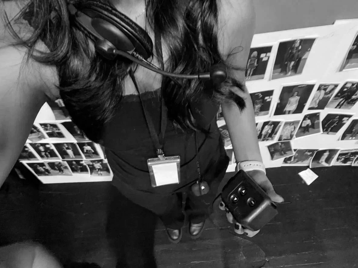 A woman with long hair wearing a headset with a microphone, holding a camera, standing in front of a display of photographs or contact sheets.