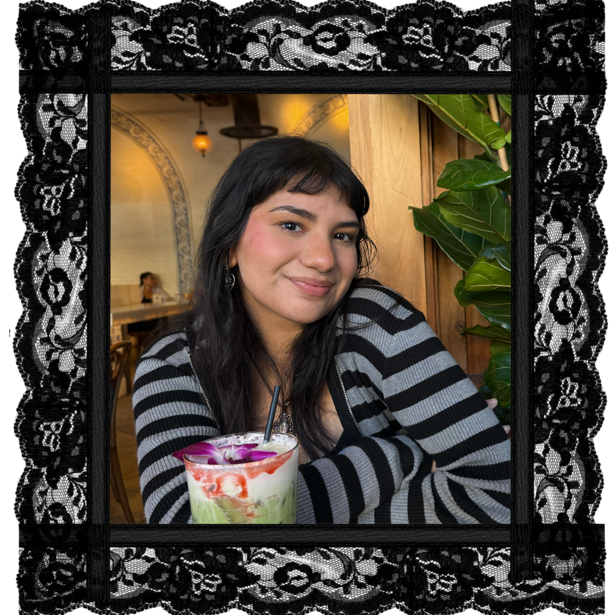 A young woman with dark hair, wearing a black and gray striped sweater, smiling at the camera inside a cozy cafe with warm lighting. She's sitting at a table with a colorful drink garnished with flowers in front of her. The background includes a wooden wall, a large green plant, and decorative lighting.