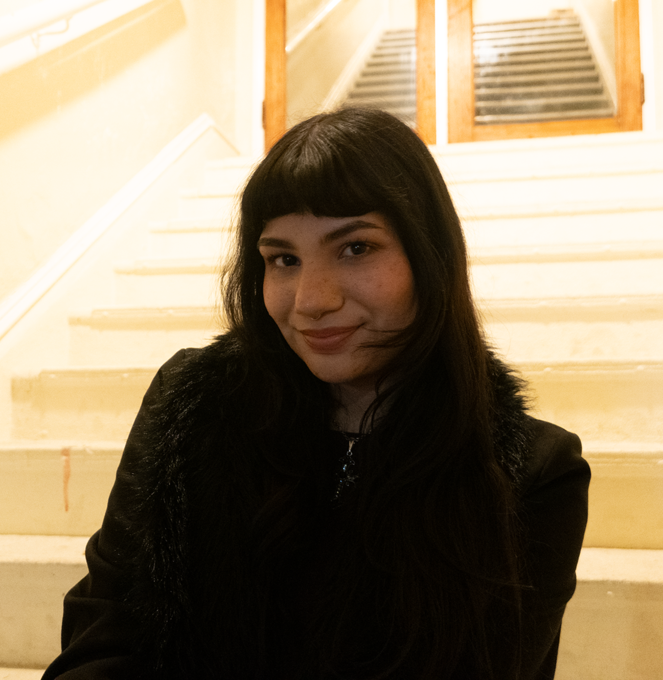A young woman with dark hair, wearing a black jacket, sitting on light-colored stairs indoors, smiling at the camera.