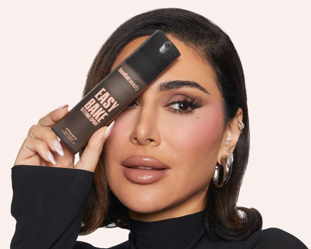 A woman with medium-length dark hair and gold jewelry holds a spray bottle labeled "Easy Bake" in front of her eye, posing against a plain background.