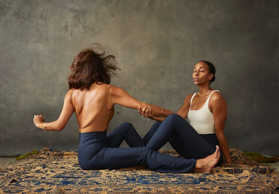 Two women are engaged in a yoga or stretching session on a patterned rug indoors, holding each other's arms and balancing in a seated position with a neutral backdrop.
