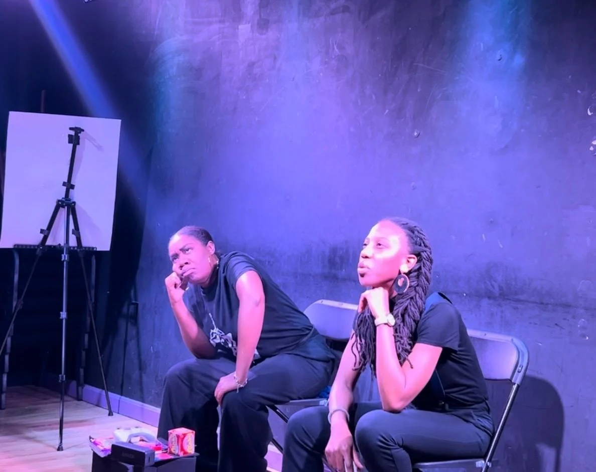 Magically Delicious in contemplation during a performance. Both Rosena and Shiyanbade are wearing black shirts and pants, are seated, and looking into the distance with their heads in their fists.