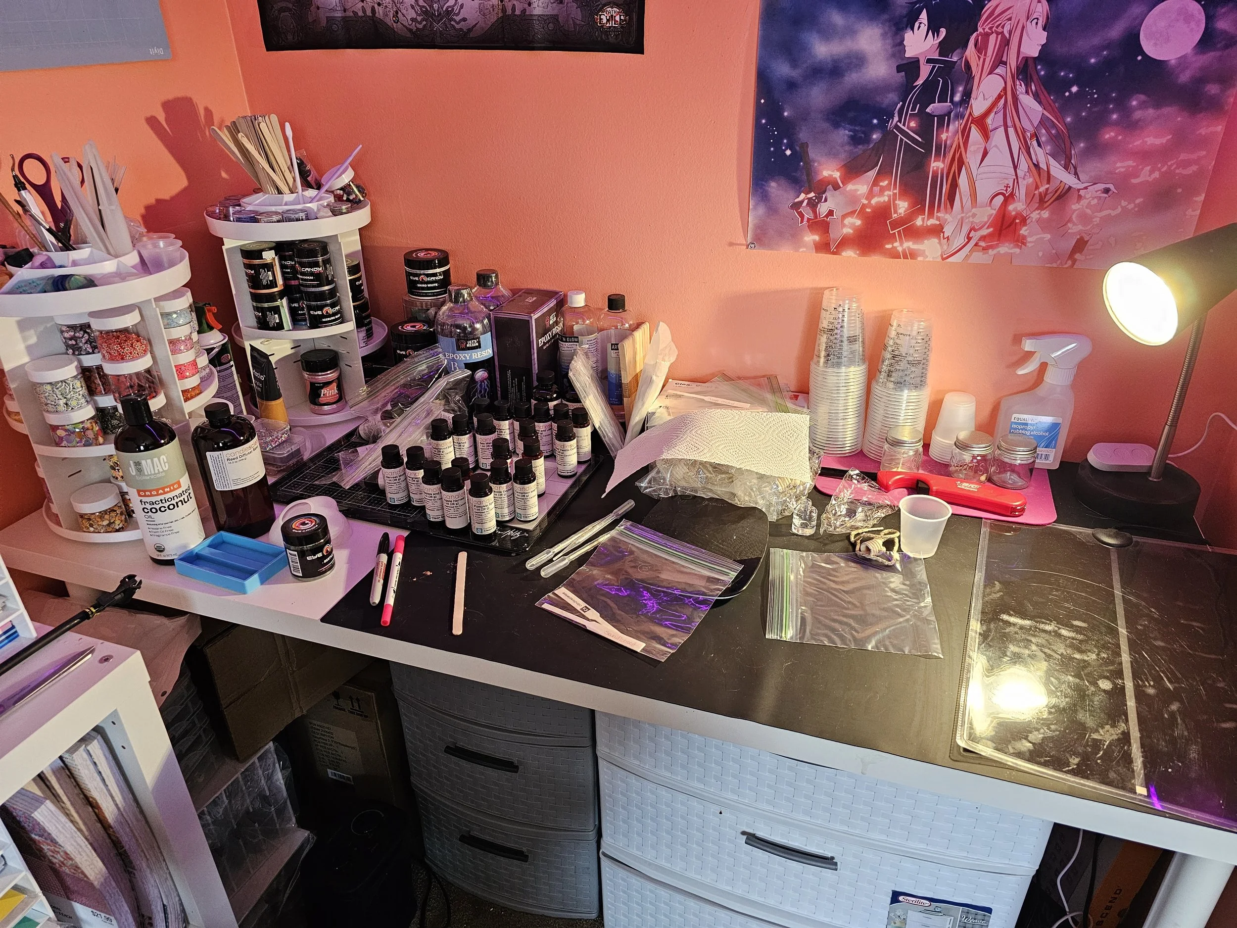 A crowded workspace with various bottles, jars, and supplies for crafting or art projects on the desk, stacked plastic cups, a lamp, and an anime poster on the wall.