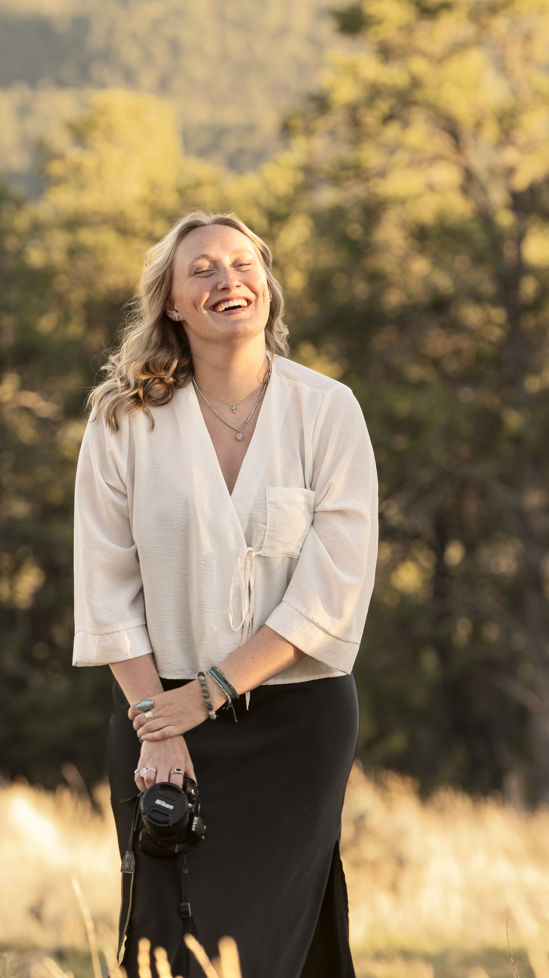 Happy woman with blonde hair, wearing a white blouse and black skirt, holding a camera, standing outdoors at sunset with trees in the background.