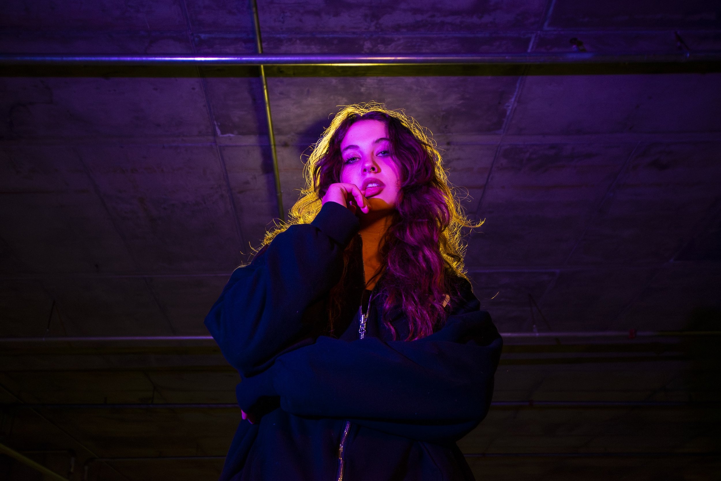 A young woman with long, wavy hair standing under a low concrete ceiling, illuminated by purple and yellow neon lights, wearing a black jacket with her hand resting on her chin.