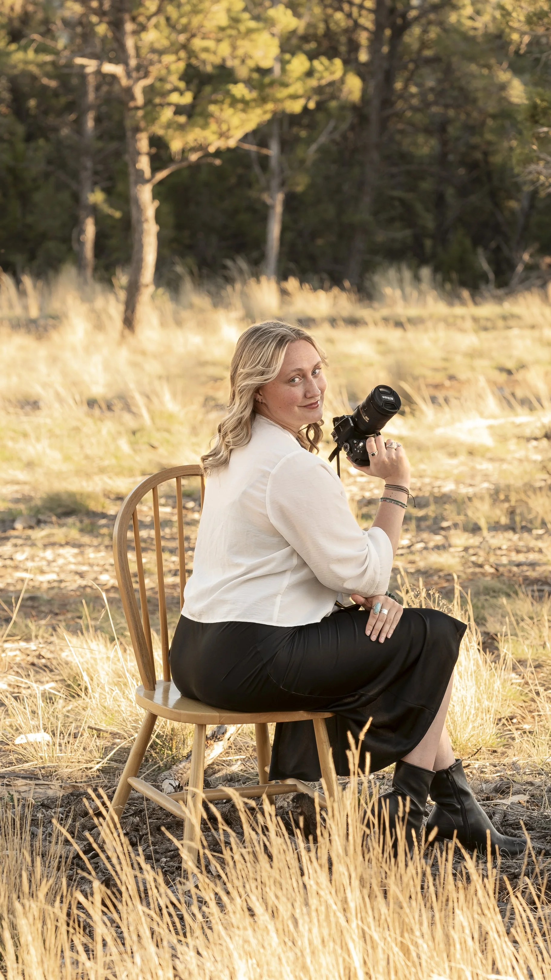 A woman with blonde hair sitting on a wooden chair outdoors in a grassy field, holding a camera, with trees in the background during sunset.